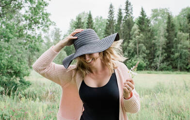 Frau läuft durch ein Feld und hält lächelnd ihren Hut - CAVF64090