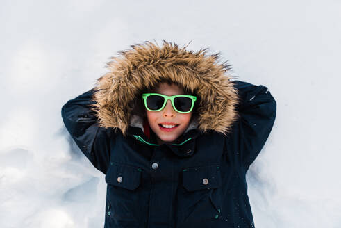 Young boy laying in snow with green sunglasses on Minnesota Spring day - CAVF64166