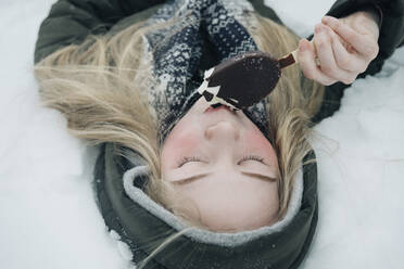 Mädchen liegt im Schnee und genießt Eiscreme - CAVF64274