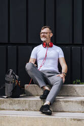 Bearded mature man sitting on steps with headphones while relaxing on a sunny day - CAVF64292