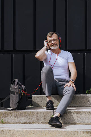 Bärtiger reifer Mann sitzt mit Kopfhörern auf den Stufen und entspannt sich an einem sonnigen Tag, lizenzfreies Stockfoto