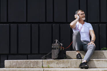 Bearded mature man sitting on steps with headphones while relaxing on a sunny day - CAVF64294