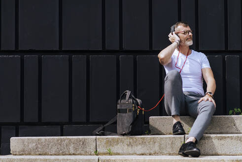 Bärtiger reifer Mann sitzt mit Kopfhörern auf den Stufen und entspannt sich an einem sonnigen Tag - CAVF64294