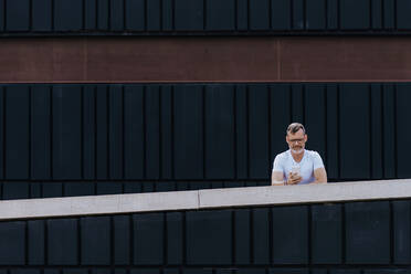 Mature bearded man using a mobile phone leaning on a fence - CAVF64299