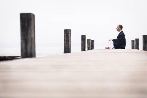 Mann sitzt auf Steg und macht Yoga-Übungen, Starnberger See, Deutschland, lizenzfreies Stockfoto