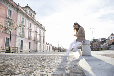 Smiling woman sitting on a bollard looking at the mobile in a street - CAVF64417