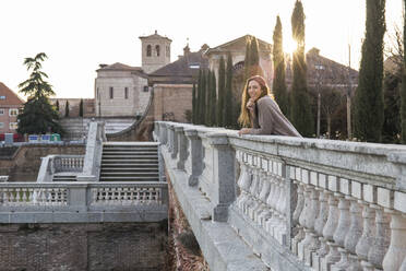 Smiling young woman leaning on a stone bridge - CAVF64427