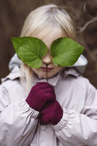 Porträt eines blonden kleinen Mädchens, das seine Augen mit grünen Blättern bedeckt - EYAF00482