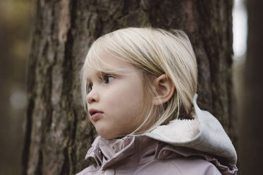 Portrait of blond little girl in front of tree trunk - EYAF00484