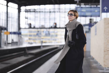 Ältere Frau mit Kamera in schwarzem Mantel und großem Wollschal wartet am Bahnsteig - EYAF00488