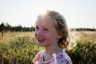 Porträt eines jungen blonden Mädchens mit lockigen Haaren, das bei Sonnenuntergang draußen lächelt - CAVF64597