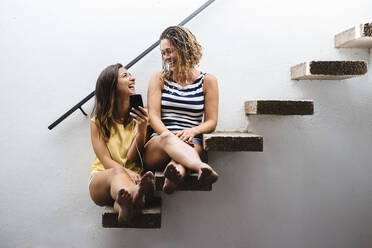 Zwei Frauen sitzen auf einer Treppe und lachen, während sie Musik hören. - CAVF64601