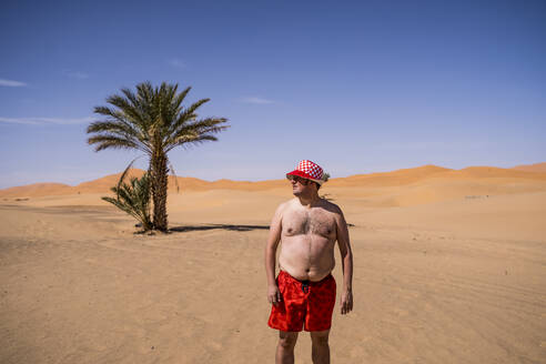 Übergewichtiger Mann mit Badeshorts und Hut in der Wüste von Marokko - OCMF00782