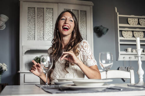 Porträt einer lachenden jungen Frau, die am gedeckten Tisch in einer Küche im Landhausstil sitzt - WFF00105