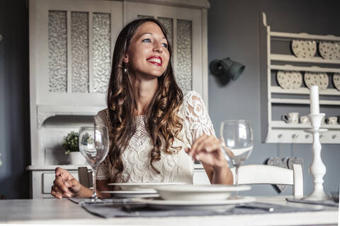 Porträt einer lächelnden jungen Frau, die an einem gedeckten Tisch in einer Küche im Landhausstil sitzt, lizenzfreies Stockfoto