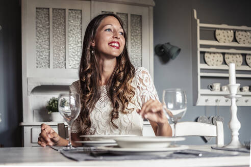 Porträt einer lächelnden jungen Frau, die an einem gedeckten Tisch in einer Küche im Landhausstil sitzt - WFF00106