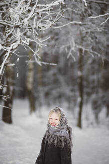 Porträt eines glücklichen kleinen Mädchens mit Kopftuch, das vor einem Winterwald steht - EYAF00496