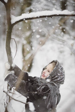 Porträt eines lächelnden kleinen Mädchens, das einen Baum im Winterwald schüttelt, lizenzfreies Stockfoto