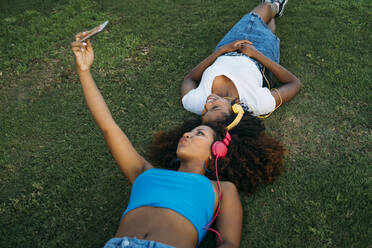 Two young women with headphones lying on a meadow taking selfie with smartphone - MPPF00086