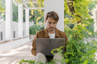 Portrait of man sitting on wall of bower using laptop - AFVF04043