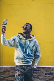 Portrait of smiling young man taking selfie with smartphone in front of yellow wall - CJMF00056