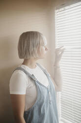 Junge Frau blickt durch die Jalousien auf das Fenster - LHPF01032