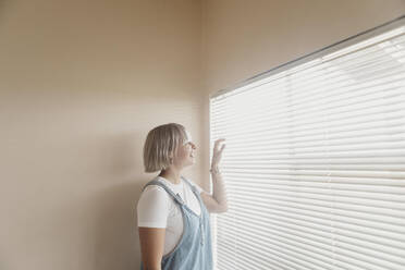 Happy young woman looking through blinds at the window - LHPF01033