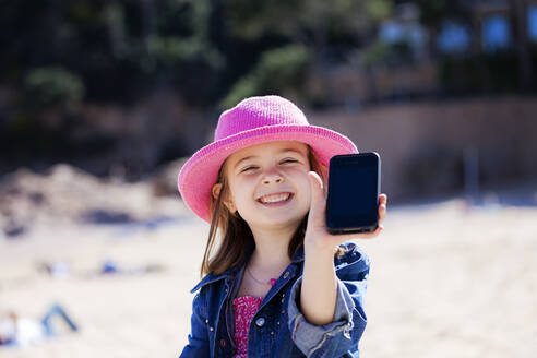 Portrait of grinning little girl showing her mobile phone on the beach - XCF00277