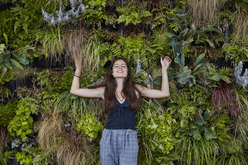 Portrait of smiling young woman with closed eyes in front of plant wall - SUF00603