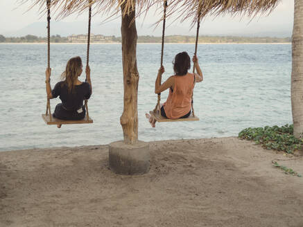 Rückenansicht von zwei Frauen, die auf Schaukeln am Meer sitzen, Gili-Inseln, Bali - KNTF03638