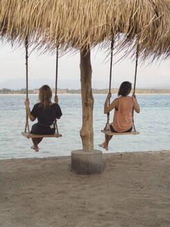 Rückenansicht von zwei Frauen, die auf Schaukeln am Meer sitzen, Gili-Inseln, Bali - KNTF03639