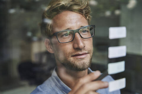 Lässiger junger Geschäftsmann mit Haftnotizen hinter einer Fensterscheibe im Büro, lizenzfreies Stockfoto