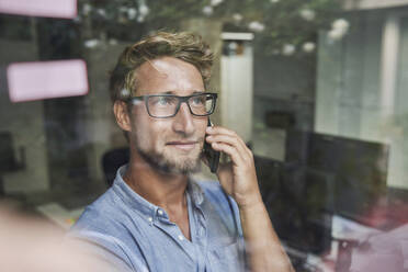 Lässiger junger Geschäftsmann am Telefon hinter einer Fensterscheibe im Büro - PNEF02089