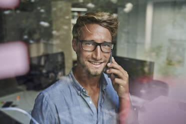 Porträt eines lächelnden jungen Geschäftsmannes am Telefon hinter einer Fensterscheibe im Büro - PNEF02090
