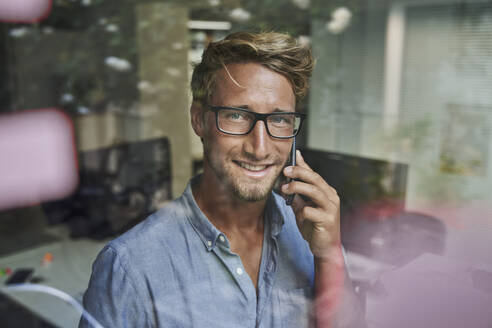 Portrait of smiling young businessman on the phone behind windowpane in office - PNEF02090