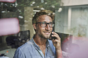 Lässiger junger Geschäftsmann am Telefon hinter einer Fensterscheibe im Büro - PNEF02091