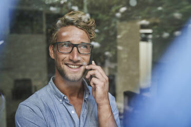 Lächelnder junger Geschäftsmann am Telefon hinter einer Fensterscheibe im Büro - PNEF02093