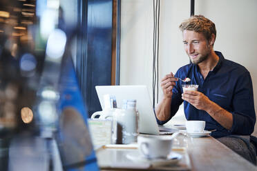 Junger Geschäftsmann isst und benutzt Laptop in einem Café - PNEF02138