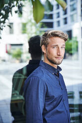 Portrait of confident young man at reflecting windowpane in the city - PNEF02154