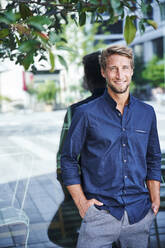 Portrait of smiling young man standing at reflecting windowpane in the city - PNEF02155