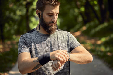 Portrait of sporty man with earphones in forest checking his smartwatch - ZEDF02642