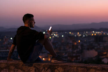 Mann schaut nachts auf sein Smartphone, die Lichter der Stadt sind im Hintergrund - CAVF64796