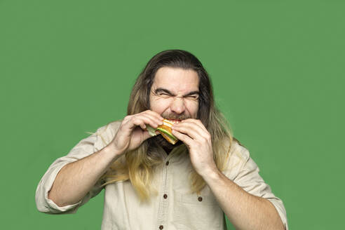 Porträt eines bärtigen Mannes, der ein Sandwich vor einem grünen Hintergrund isst - VGF00309