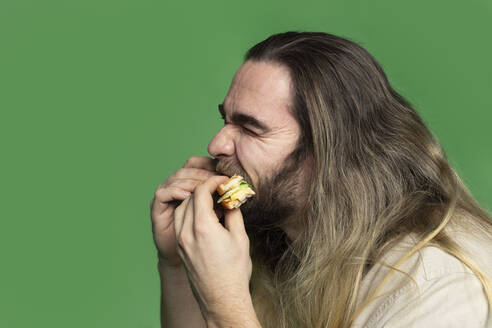 Bearded man eating sandwich in front of green background - VGF00310