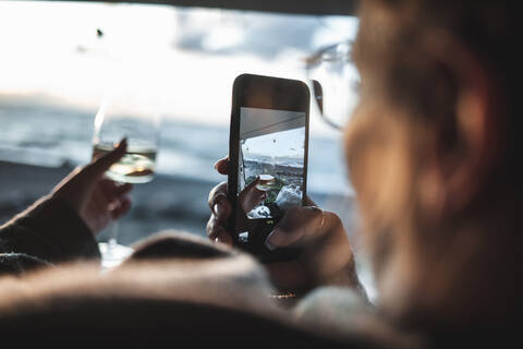 Frau, die ein Glas Weißwein mit ihrem Smartphone am Strand fotografiert, lizenzfreies Stockfoto