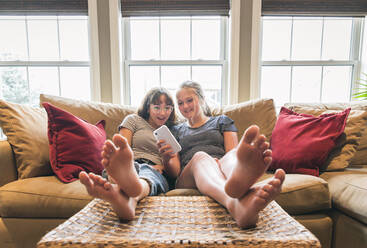 Zwei Mädchen im Teenageralter sitzen mit hochgelegten Füßen auf der Couch und schauen auf ihr Handy. - CAVF64814