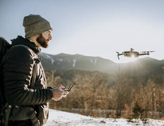 Ein Mann mit Bart fliegt im Winter eine Drohne in den Bergen. - CAVF64830