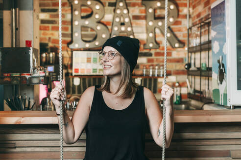 Smiling woman sitting on a swing in a hotel bar - NAF00149