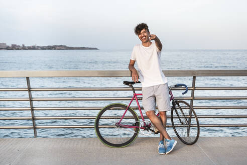 Junger Mann mit Fahrrad, stehend auf Brücke am Meer - RCPF00036