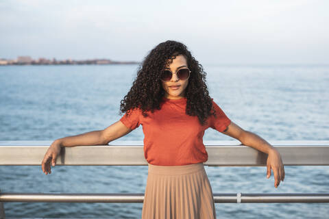 Schöne Frau mit Sonnenbrille, die sich am Meer an ein Geländer lehnt, lizenzfreies Stockfoto